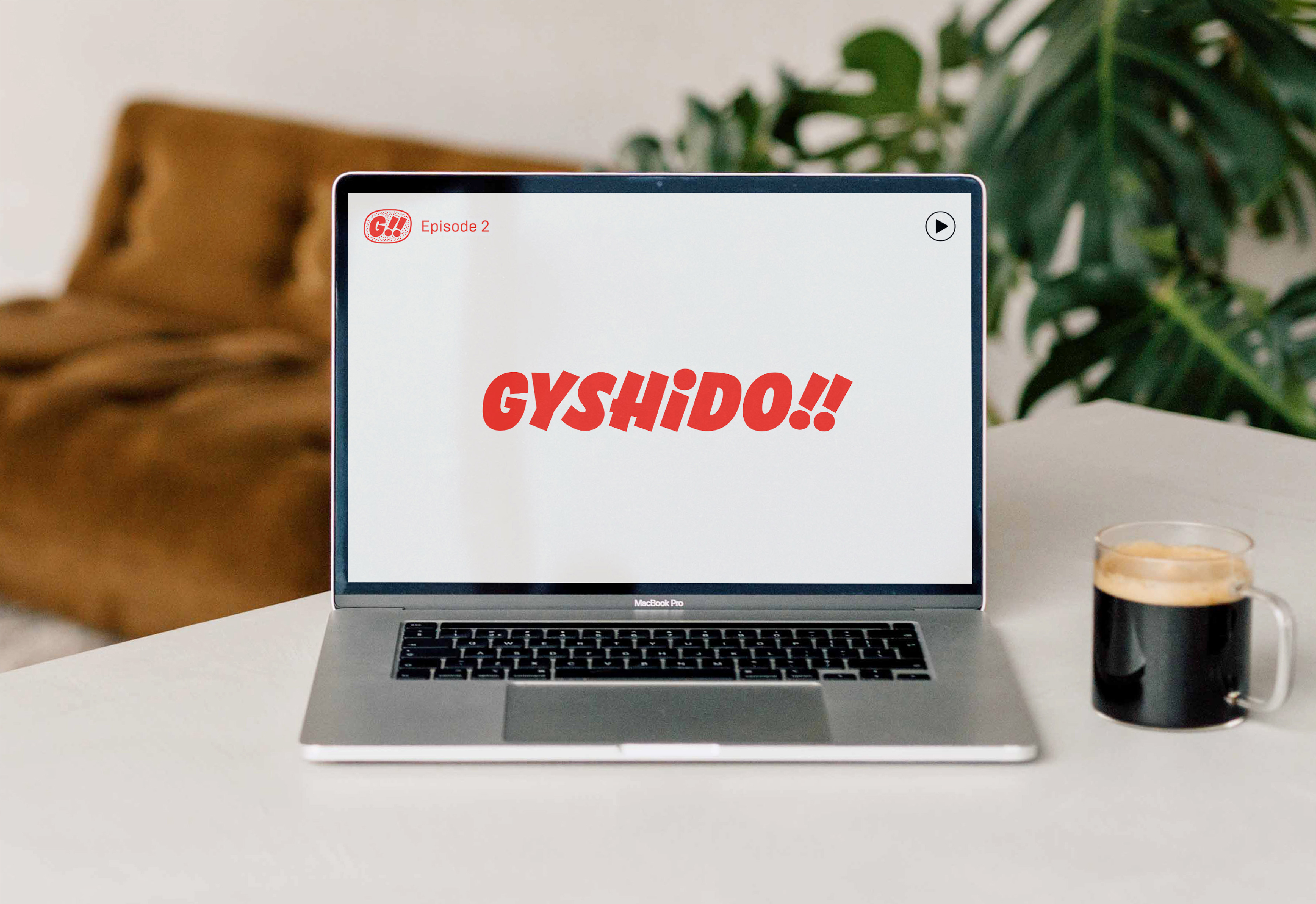 GYSHIDO!! red logotype and G!! badge shown on laptop screen in lifestyle mockup with coffee and plants